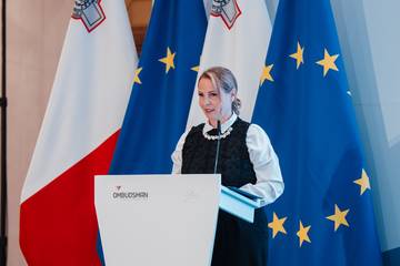 Namestnica varuha dr. Dijana Možina Zupanc za govorniškim pultom. V ozadju so zastave. 